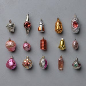 May include: A collection of vintage Christmas ornaments in various shapes and colors, including silver, gold, red, and pink. The ornaments are arranged on a gray background, showcasing their diverse designs and textures.