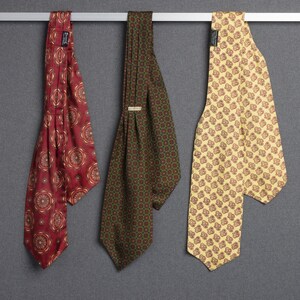 May include: Three neckties hanging side by side. The left tie is deep red with a repeating circular medallion pattern. The center tie is dark green with a small geometric grid print. The right tie is pale yellow with a repeating brown floral motif.