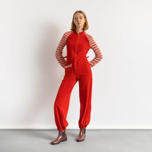 May include: A full-length, red jumpsuit with a zip-up front and long sleeves. The sleeves have a patterned design in red, white, and beige. The jumpsuit has pockets and is paired with brown ankle boots. The outfit is a vintage style.