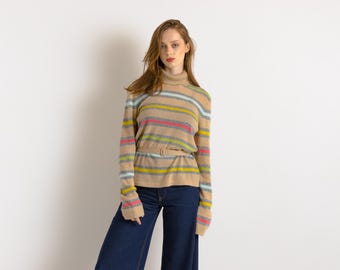 Puede incluir: Un jersey de cuello alto a rayas con base marrón y rayas de colores. El jersey tiene un cinturón en la cintura y se lleva con vaqueros azules.