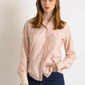 Puede incluir: Una mujer con una camisa de manga larga a rayas con botones en tonos melocot&oacute;n, blanco y azul. Lleva pantalones vaqueros azul oscuro y tiene la mano en el bolsillo.