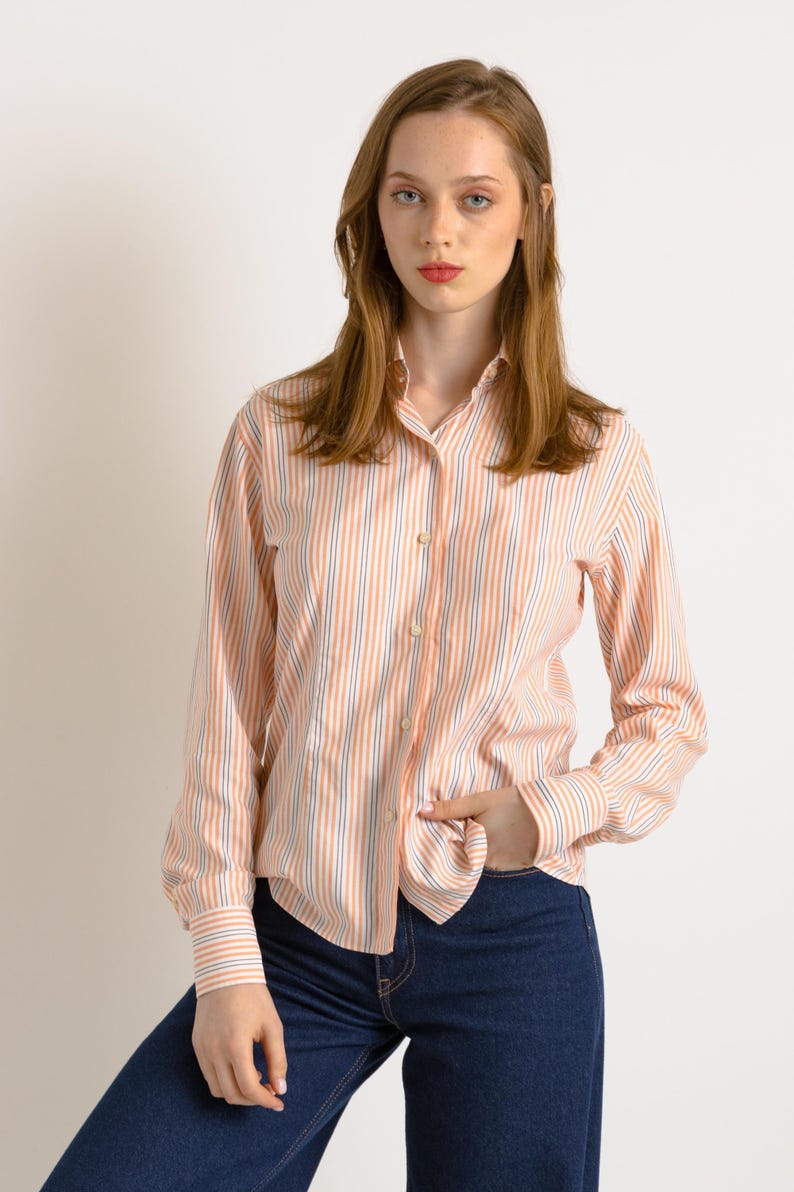 Puede incluir: Una mujer con una camisa de manga larga a rayas con botones en tonos melocot&oacute;n, blanco y azul. Lleva pantalones vaqueros azul oscuro y tiene la mano en el bolsillo.