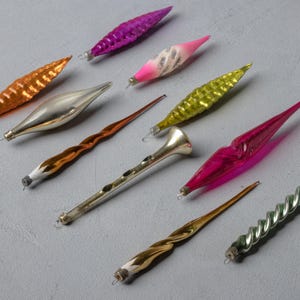 Puede incluir: Una colección de adornos navideños vintage en varias formas y colores. Los adornos incluyen formas de lágrima, espiral y trompeta en tonos dorados, plateados, rosas, morados y verdes. Cada adorno tiene una pequeña tapa de metal para colgar.