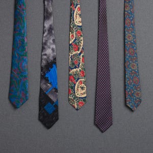 Puede incluir: Cinco corbatas estampadas de varios colores cuelgan sobre un fondo gris. Las corbatas presentan diseños de cachemira, florales y geométricos en tonos de azul, rojo y morado. Una corbata es morada lisa con lunares blancos.