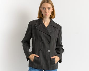 Puede incluir: Una blazer cruzada gris oscuro con finas rayas blancas. La blazer tiene dos filas de botones negros y dos bolsillos delanteros. La modelo lleva vaqueros azules.