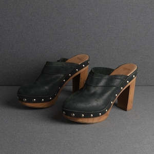 May include: A pair of black leather clogs with wooden platform soles and heels. The clogs have silver-colored stud accents around the soles and straps. The heels are approximately 4 inches high.