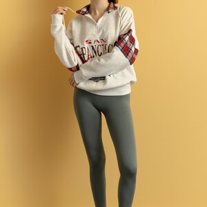 May include: A woman wearing a white sweatshirt with a red and black plaid sleeve and the text "SAN FRANCISCO" on the front. She is also wearing gray leggings and red sandals.