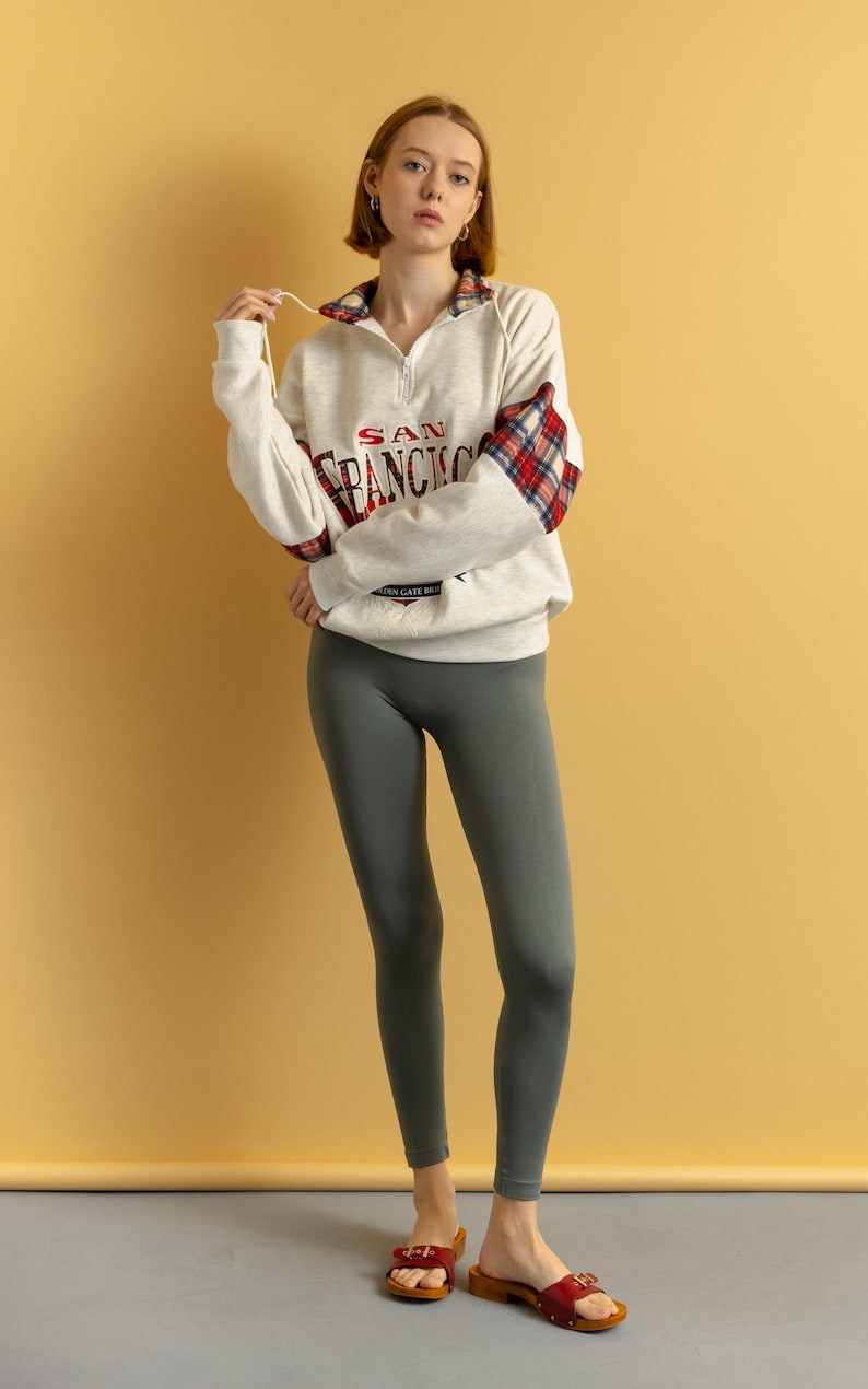 May include: A woman wearing a white sweatshirt with a red and black plaid sleeve and the text "SAN FRANCISCO" on the front. She is also wearing gray leggings and red sandals.