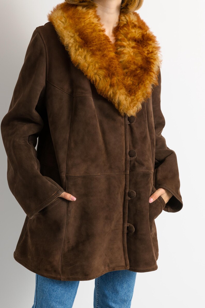 May include: A brown suede coat with a large, fluffy, tan-colored collar. The coat has a button-up front, pockets, and a classic design. The coat is a medium length and appears to be vintage.