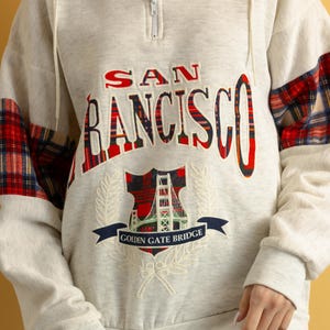 May include: A white sweatshirt with a red and black plaid design on the sleeves. The sweatshirt has a zipper closure and features an embroidered graphic of the Golden Gate Bridge with the text "SAN FRANCISCO GOLDEN GATE BRIDGE".