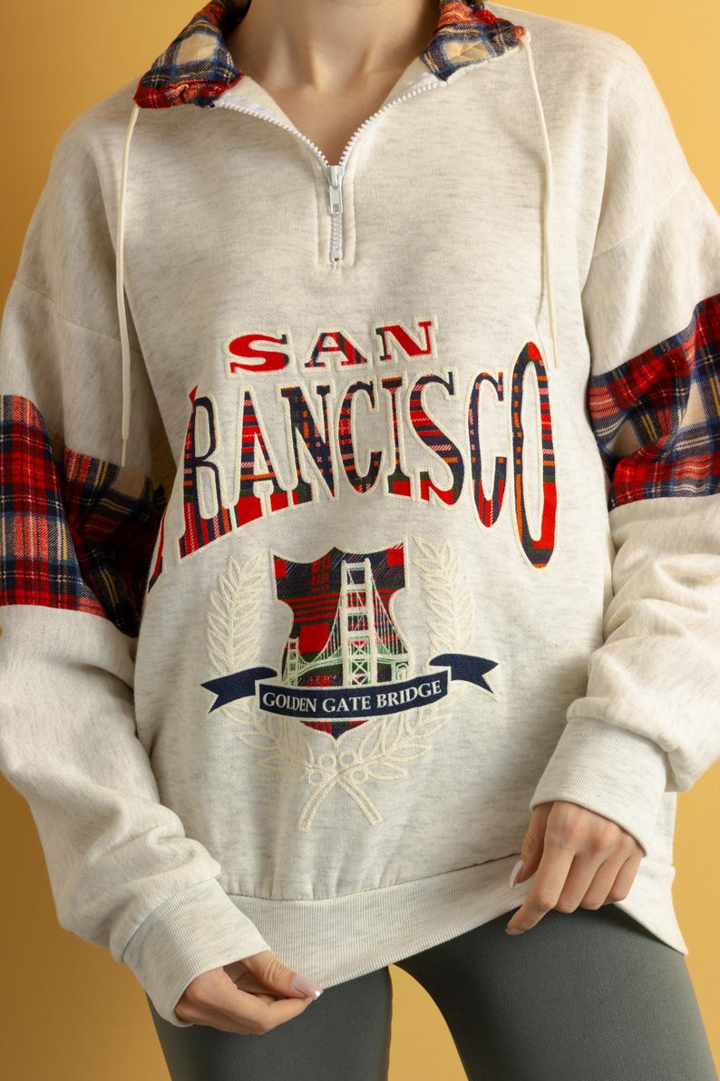 May include: A white sweatshirt with a red and black plaid design on the sleeves. The sweatshirt has a zipper closure and features an embroidered graphic of the Golden Gate Bridge with the text "SAN FRANCISCO GOLDEN GATE BRIDGE".