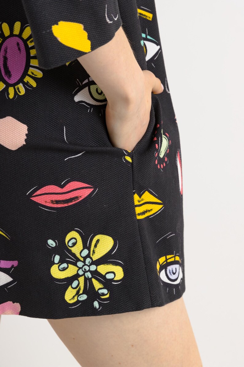 May include: A black dress with a colorful print of eyes, lips, and flowers. The dress has a pocket on the side.