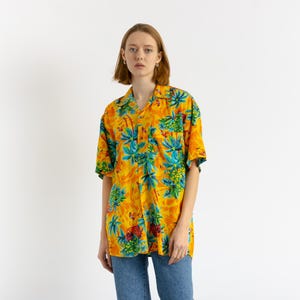 Könnte beinhalten: Ein gelbes, kurzärmeliges Hemd mit tropischem Muster, das Palmen, Blumen und Ananas zeigt. Das Hemd hat einen Knopfkrag und eine Tasche auf der linken Seite.