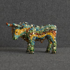 May include: A bull figurine covered in a colorful mosaic pattern of small, irregular tiles in shades of turquoise, yellow, green, brown, and white. The sculpture features a standing posture with horns, a defined snout, and four legs.