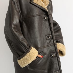 May include: A dark brown leather coat with a beige shearling lining. The coat features a notched collar, button closures, and shearling trim on the cuffs and collar. A hand is in the pocket.