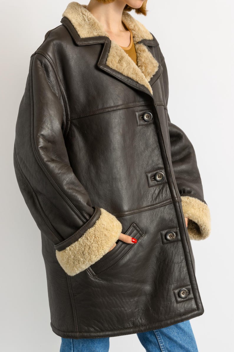 May include: A dark brown leather coat with a beige shearling lining. The coat features a notched collar, button closures, and shearling trim on the cuffs and collar. A hand is in the pocket.