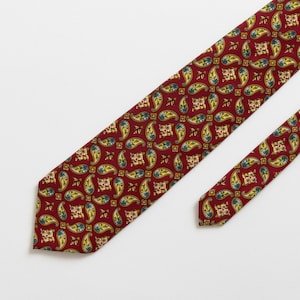 Può includere: Una cravatta rossa con un motivo paisley ripetuto in giallo, blu e crema. La cravatta è angolata su uno sfondo bianco, evidenziando il suo design e la sua consistenza. È presente anche una cravatta sottile abbinata.