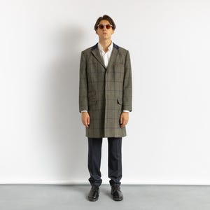 May include: A long, olive green and brown plaid coat with a navy blue collar, worn over a white shirt and dark blue pants. The coat has two front pockets and a classic, tailored design. The person is wearing black shoes and sunglasses.
