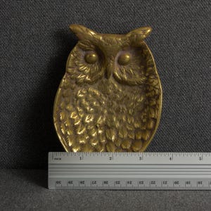 May include: A gold-toned metal owl-shaped trinket dish with detailed feather textures and prominent eyes. A metal ruler is positioned at the base, showing the object measures approximately 4.5 inches in width at its widest point.