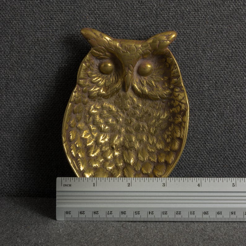 May include: A gold-toned metal owl-shaped trinket dish with detailed feather textures and prominent eyes. A metal ruler is positioned at the base, showing the object measures approximately 4.5 inches in width at its widest point.