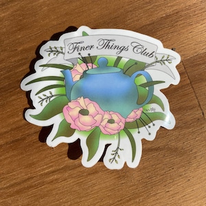 Puede incluir: Una pegatina blanca con una tetera azul rodeada de hojas verdes y flores rosas. El texto "Finer Things Club" está escrito en una pancarta sobre la tetera.