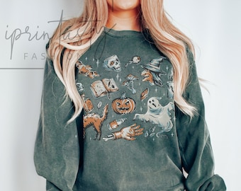 May include: A green long-sleeved sweatshirt with a vintage-style Halloween graphic print featuring a witch, a ghost, a pumpkin, a cat, and other spooky elements. The text "BLUE SPRUCE" is visible on the bottom right corner of the image.