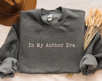 May include: Dark grey sweatshirt with the text "In My Author Era." A brown hat and dried flowers are next to the sweatshirt. The sweatshirt has long sleeves and a crew neck.