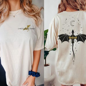 Puede incluir: Una camiseta blanca con un gr&aacute;fico de drag&oacute;n dorado y el texto "I will not die today" en la parte delantera. La parte posterior de la camiseta presenta un gr&aacute;fico de drag&oacute;n negro con una luna creciente y estrellas.