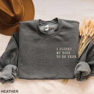 May include: Dark heather grey sweatshirt with the text "I closed my book to be here." printed in white.