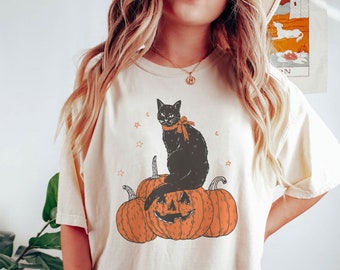 May include: A cream colored t-shirt with a black cat sitting on top of a jack-o-lantern. The cat is wearing a red bow. The t-shirt has the words "iprinttasty" and "FASHION" printed on the bottom left corner. The t-shirt is labeled "Ivory".