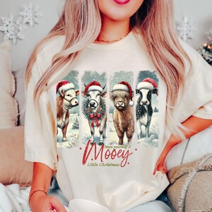 May include: Ivory-colored t-shirt featuring a festive design. The graphic showcases four cows wearing Santa hats, set against a snowy backdrop. Text reads "Have yourself a Mooey Little Christmas" in red script.