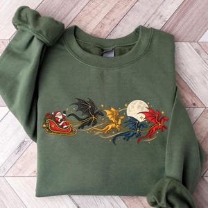May include: An olive green crewneck sweatshirt featuring a graphic print of Santa Claus in a red sleigh pulled by four flying dragons in black, gold, blue, and red colors, set against a full moon and small yellow stars.