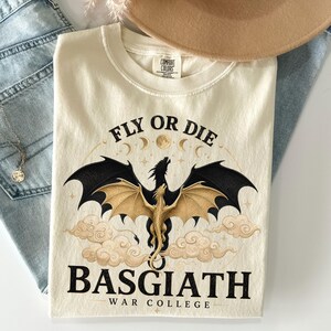May include: Cream-coloured t-shirt featuring a dragon graphic and the text "FLY OR DIE" and "BASGIATH WAR COLLEGE". The dragon has black and gold wings. The shirt is made of a soft material.