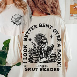 May include: A white t-shirt with a black graphic print of a skeleton woman reading a book. The text on the shirt reads "I look better bent over a book, Proud Smut Reader".
