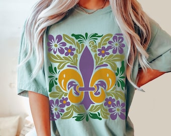 May include: A light green t-shirt with a purple and gold fleur-de-lis design surrounded by green leaves and purple flowers. The text "BAY" is visible in the bottom right corner of the image.