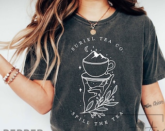 May include: A dark gray t-shirt with a white graphic design of a hand holding a teacup with a mountain inside. The text "Suriel Tea Co." is above the graphic and "Spill the tea" is below.