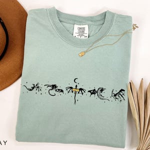 May include: A light green t-shirt with a crew neck and short sleeves. The shirt features a black dragon graphic design across the chest. A gold necklace with a small pendant is draped on the shirt.