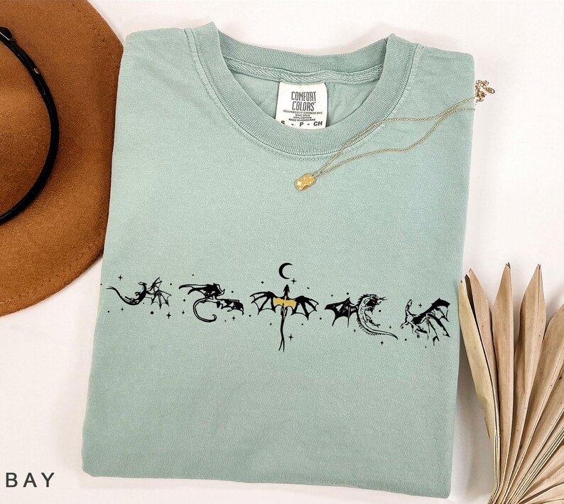 May include: A light green t-shirt with a crew neck and short sleeves. The shirt features a black dragon graphic design across the chest. A gold necklace with a small pendant is draped on the shirt.