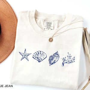 Op de afbeelding: Een wit T-shirt met een blauw ontwerp van een zeester, schelpen en zeewier. Het shirt heeft de tekst "Comfort Colors" en "S - P - CN" op de voorkant.
