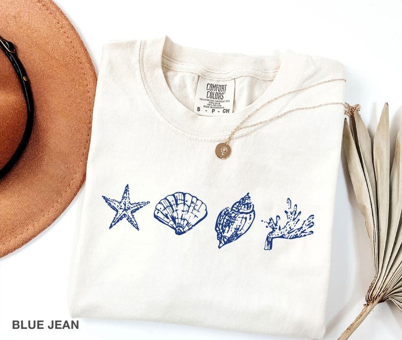 Op de afbeelding: Een wit T-shirt met een blauw ontwerp van een zeester, schelpen en zeewier. Het shirt heeft de tekst "Comfort Colors" en "S - P - CN" op de voorkant.