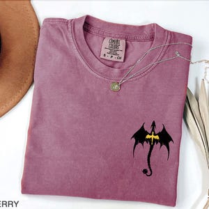 May include: A mauve colored t-shirt with a black dragon graphic and a gold chain necklace. The t-shirt has the text "COMFORT COLORS" on the tag.