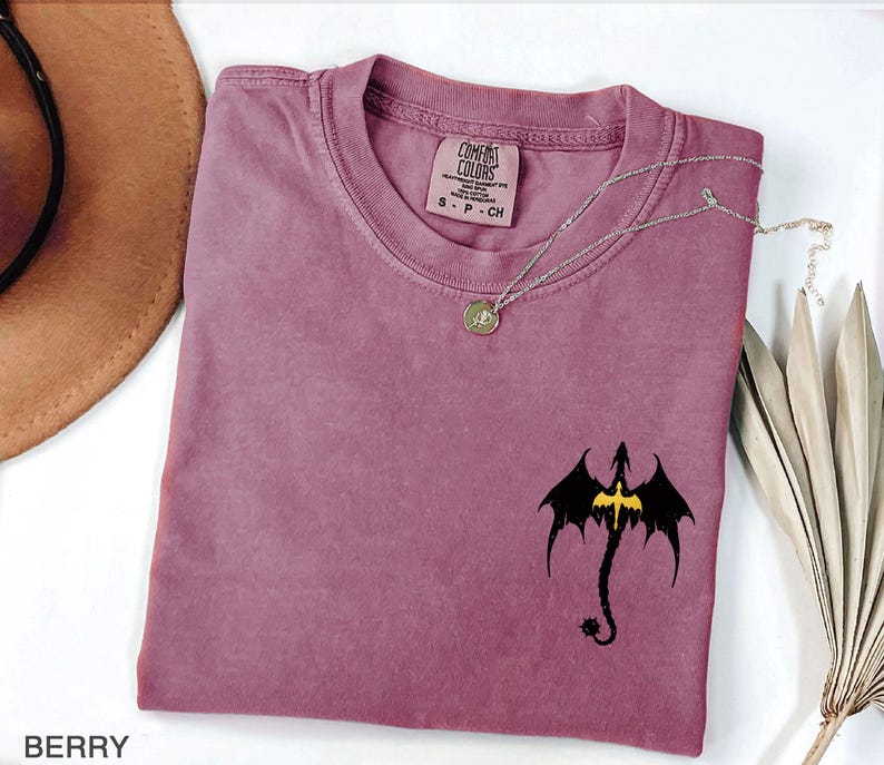 May include: A mauve colored t-shirt with a black dragon graphic and a gold chain necklace. The t-shirt has the text "COMFORT COLORS" on the tag.