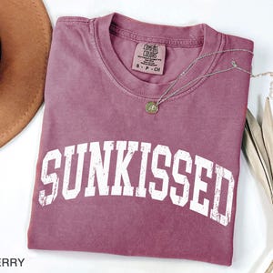 May include: Berry-colored t-shirt with the word "SUNKISSED" in white block letters. A silver necklace with a pendant rests on the shirt. The shirt is folded on a white surface, with a brown hat and dried plant in the background.