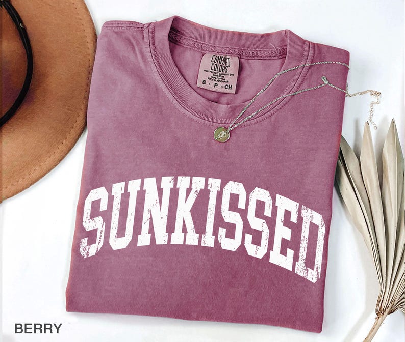 May include: Berry-colored t-shirt with the word "SUNKISSED" in white block letters. A silver necklace with a pendant rests on the shirt. The shirt is folded on a white surface, with a brown hat and dried plant in the background.