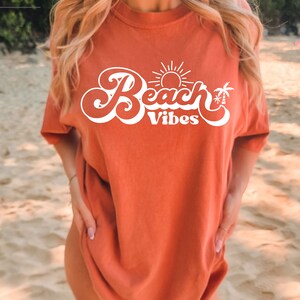 May include: A woman wearing a burnt orange t-shirt with a white graphic that says "Beach Vibes" with a sun and palm trees.