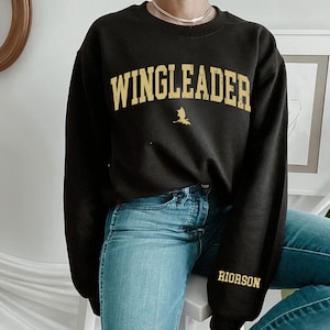 May include: Black sweatshirt with the text "WINGLEADER" in gold lettering and a small gold dragon graphic. The sweatshirt also has the text "RIORSON" in gold lettering on the left sleeve.