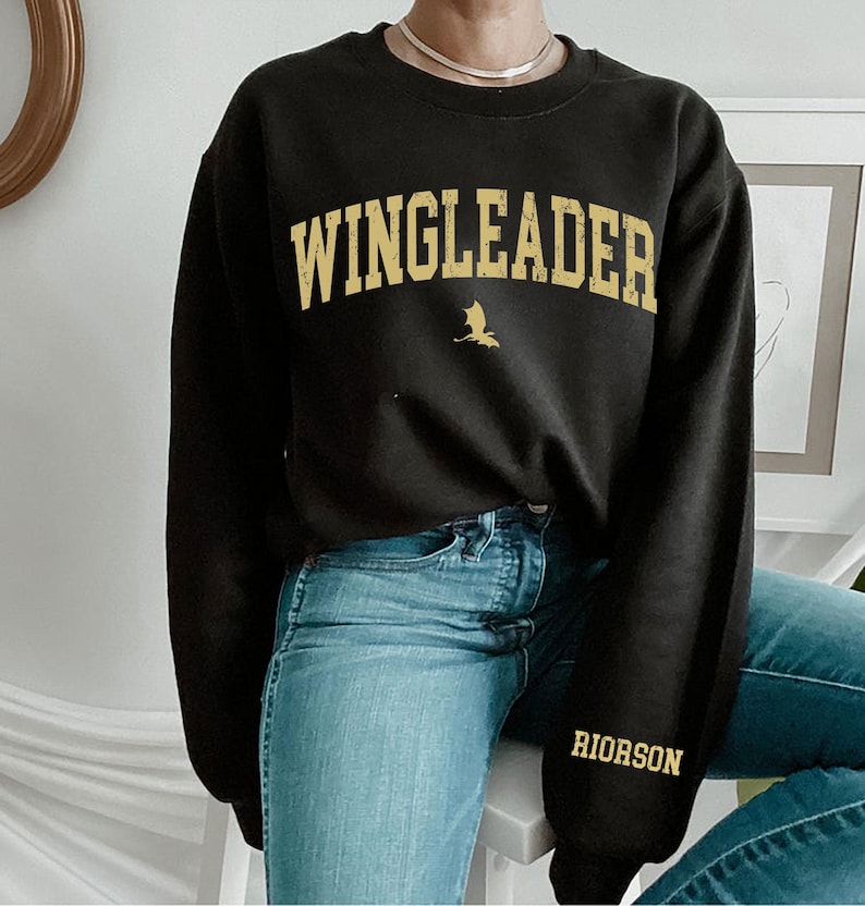 May include: Black sweatshirt with the text "WINGLEADER" in gold lettering and a small gold dragon graphic. The sweatshirt also has the text "RIORSON" in gold lettering on the left sleeve.