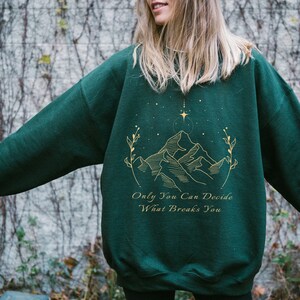 May include: A dark green sweatshirt with a gold mountain range design and the text "Only You Can Decide What Breaks You".