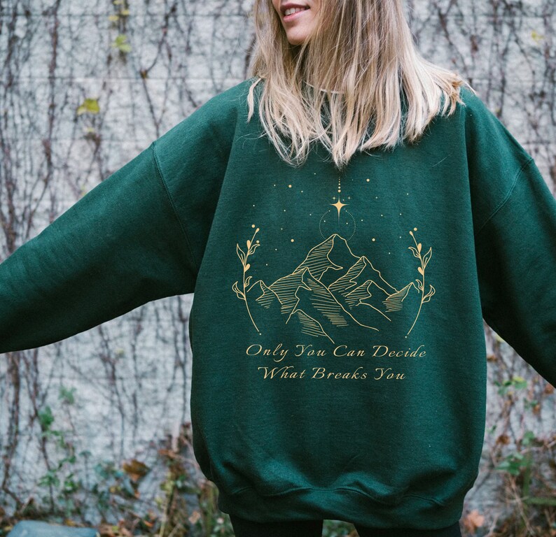 May include: A dark green sweatshirt with a gold mountain range design and the text "Only You Can Decide What Breaks You".