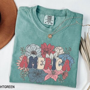 May include: Light green t-shirt with a floral design and the word "AMERICA" in colorful, retro-style lettering. The shirt is made of a soft, comfortable material and has a relaxed fit.